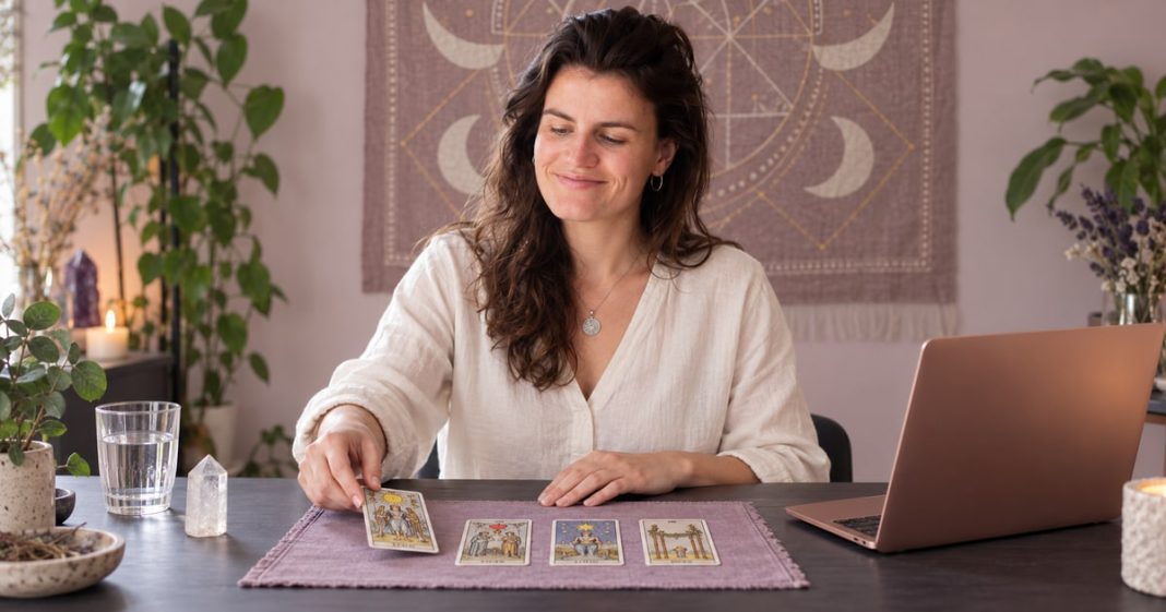 A psychic reader laying a tarot card on a lilac mat beside a correctly positioned laptop and quartz.
